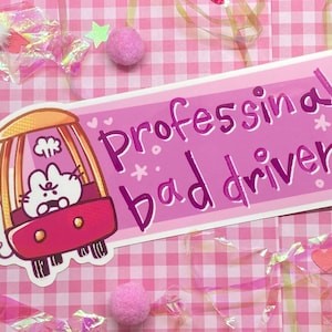 May include: A pink sticker with the text "Professional bad driver" in a playful font. The sticker features a cartoon car with a bunny driver. The background is a pink gingham pattern with decorative elements.