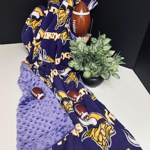 Puede incluir: Una manta de forro polar morada y dorada con el logotipo y el texto de los Minnesota Vikings. La manta está drapeada sobre una superficie blanca, con una tela morada con hoyuelos en la parte inferior. Varias miniaturas de fútbol están colocadas sobre la manta.