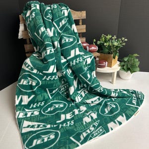 Puede incluir: Una manta de forro polar verde y blanco con el texto "JETS" y otros gráficos del equipo. La manta está doblada sobre una silla de madera. La manta está hecha de un material suave.