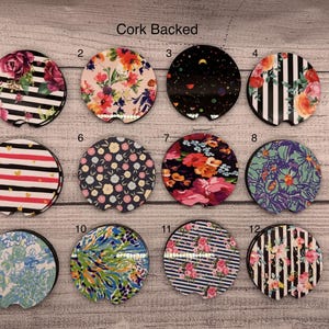 May include: A collection of twelve cork-backed car coasters with various floral, striped, and patterned designs. The coasters feature a small notch for easy removal and are arranged on a wooden surface. The text "Cork Backed" is visible.