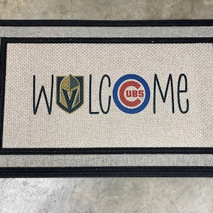 May include: A welcome mat with a black border and a white background. The word "WELCOME" is written in black with the Vegas Golden Knights logo on the left and the Chicago Cubs logo on the right.
