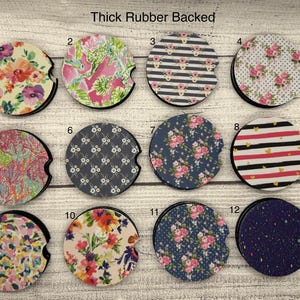 May include: A collection of twelve car coasters with various designs. The coasters feature floral, striped, and patterned prints in colors like pink, blue, and yellow. Each coaster has a black rubber backing and a cutout for easy removal.