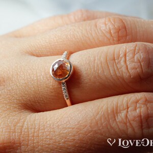 Pu&ograve; includere: Un anello in oro rosa con una pietra preziosa rotonda marrone e una fascia di piccole pietre preziose bianche.