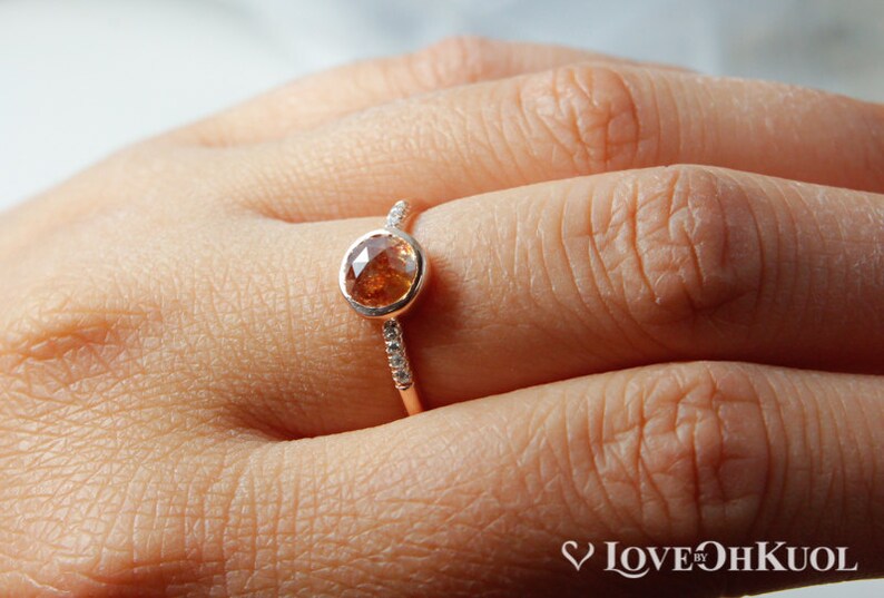 Pu&ograve; includere: Un anello in oro rosa con una pietra preziosa rotonda marrone e una fascia di piccole pietre preziose bianche.