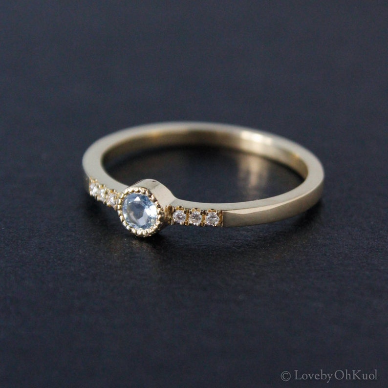 May include: A delicate gold band ring with a small, light blue gemstone set in a bezel setting. The band is accented with a row of tiny diamonds.