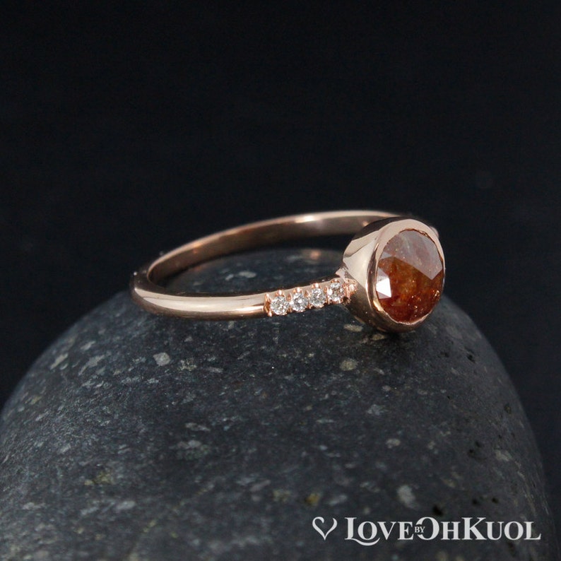 Pu&ograve; includere: Un anello in oro rosa con una pietra rotonda marrone e una fila di piccoli diamanti.