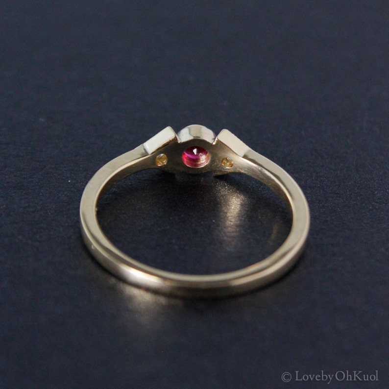 May include: A gold ring with a red gemstone and two smaller white gemstones. The ring is on a black background.