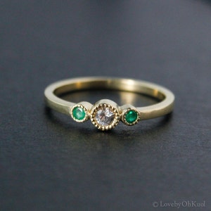 K&ouml;nnte beinhalten: Ein goldener Ring mit einem runden wei&szlig;en Edelstein und zwei kleineren gr&uuml;nen Edelsteinen auf jeder Seite.