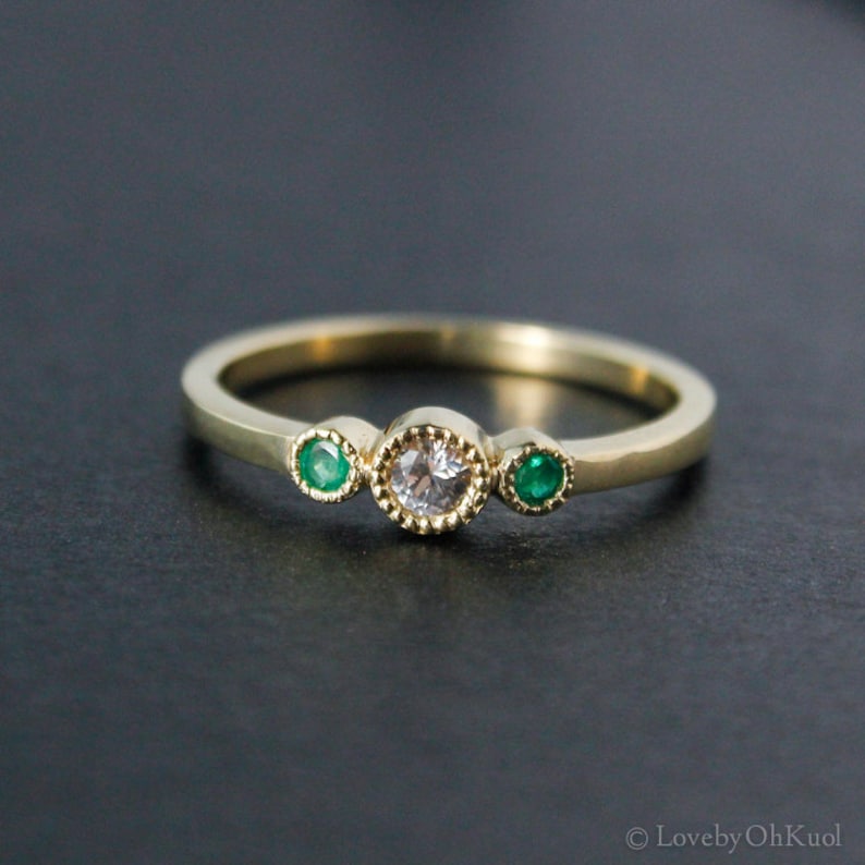 K&ouml;nnte beinhalten: Ein goldener Ring mit einem runden wei&szlig;en Edelstein und zwei kleineren gr&uuml;nen Edelsteinen auf jeder Seite.