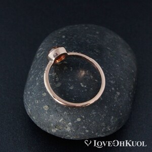 Pu&ograve; includere: Un anello in oro rosa con una pietra preziosa rotonda di colore arancione incastonata in una montatura a castone. L'anello &egrave; su una pietra grigia.