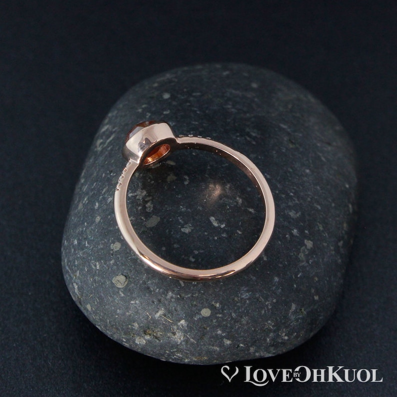Pu&ograve; includere: Un anello in oro rosa con una pietra preziosa rotonda di colore arancione incastonata in una montatura a castone. L'anello &egrave; su una pietra grigia.
