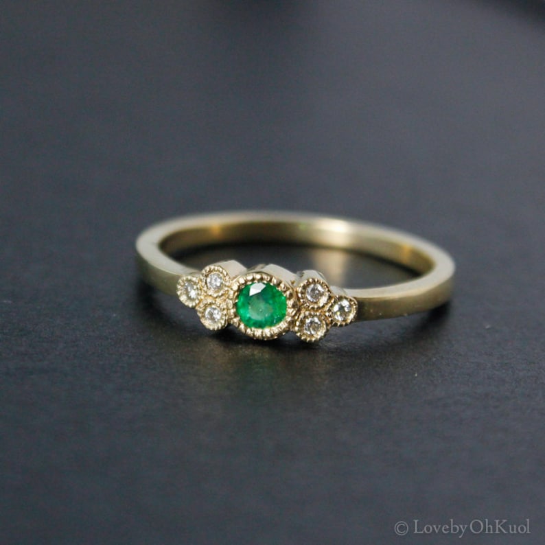 Pu&ograve; includere: Un anello d'oro con uno smeraldo verde circondato da diamanti pi&ugrave; piccoli.