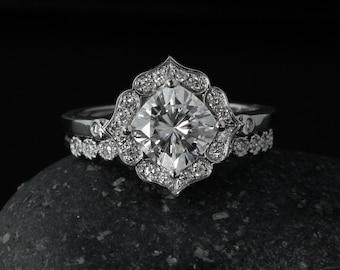 Puede incluir: Un anillo de compromiso de plata con una gran piedra central redonda y clara rodeada de piedras más pequeñas. El anillo tiene un diseño floral delicado.