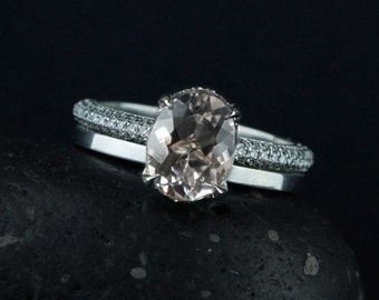 Op de afbeelding: Een delicate verlovingsring met een grote ovale morganiet-edelsteen, omringd door een halo van kleinere diamanten. De ring is gezet in een witgouden band en wordt geaccentueerd door een bijpassende diamantband.