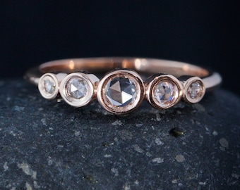 May include: A rose gold band with five bezel-set round diamonds. The diamonds are all different sizes and have a slightly cloudy appearance.