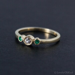 K&ouml;nnte beinhalten: Ein goldener Ring mit einem einzigen runden wei&szlig;en Edelstein und zwei kleineren runden gr&uuml;nen Edelsteinen.