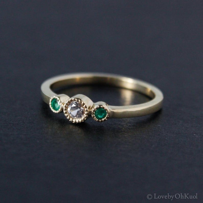 K&ouml;nnte beinhalten: Ein goldener Ring mit einem einzigen runden wei&szlig;en Edelstein und zwei kleineren runden gr&uuml;nen Edelsteinen.