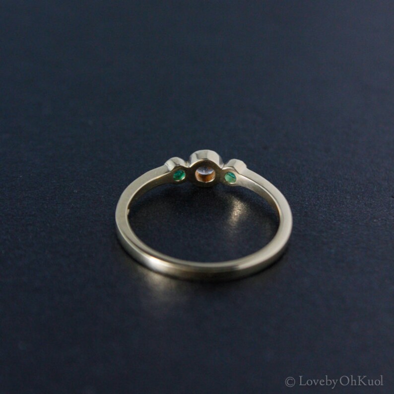 K&ouml;nnte beinhalten: Ein goldener Ring mit einem runden, klaren Mittelst&uuml;ck und zwei kleineren gr&uuml;nen Steinen auf jeder Seite.