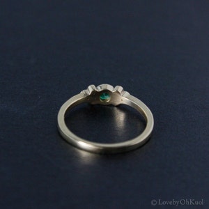 Pu&ograve; includere: Un anello d'oro con una pietra preziosa verde e due pietre bianche pi&ugrave; piccole. L'anello &egrave; su uno sfondo scuro.