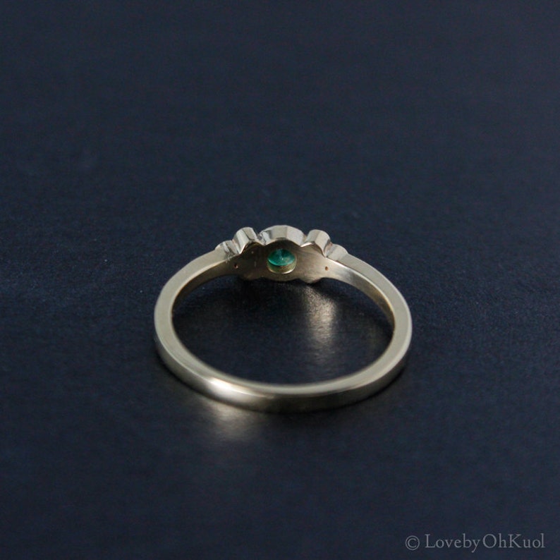 Pu&ograve; includere: Un anello d'oro con una pietra preziosa verde e due pietre bianche pi&ugrave; piccole. L'anello &egrave; su uno sfondo scuro.