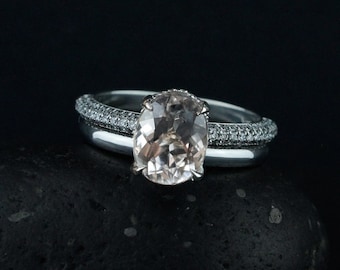 Op de afbeelding: Een zilveren ring met een grote ovale roze edelsteen, omringd door kleinere diamanten. De ring is geplaatst op een zwarte achtergrond.