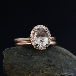 May include: A rose gold engagement ring with a large oval-shaped center stone surrounded by a halo of smaller diamonds. The ring is set on a thin band and is accompanied by a matching wedding band.
