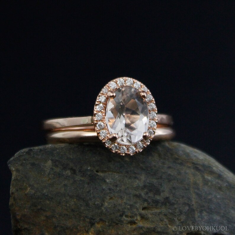 May include: A rose gold engagement ring with a large oval-shaped center stone surrounded by a halo of smaller diamonds. The ring is set on a thin band and is accompanied by a matching wedding band.