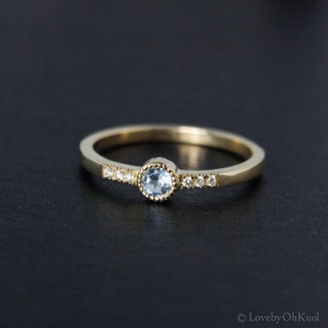 May include: A delicate gold band ring with a small, round, light blue gemstone set in a bezel setting. The band is accented with a row of tiny, clear gemstones.