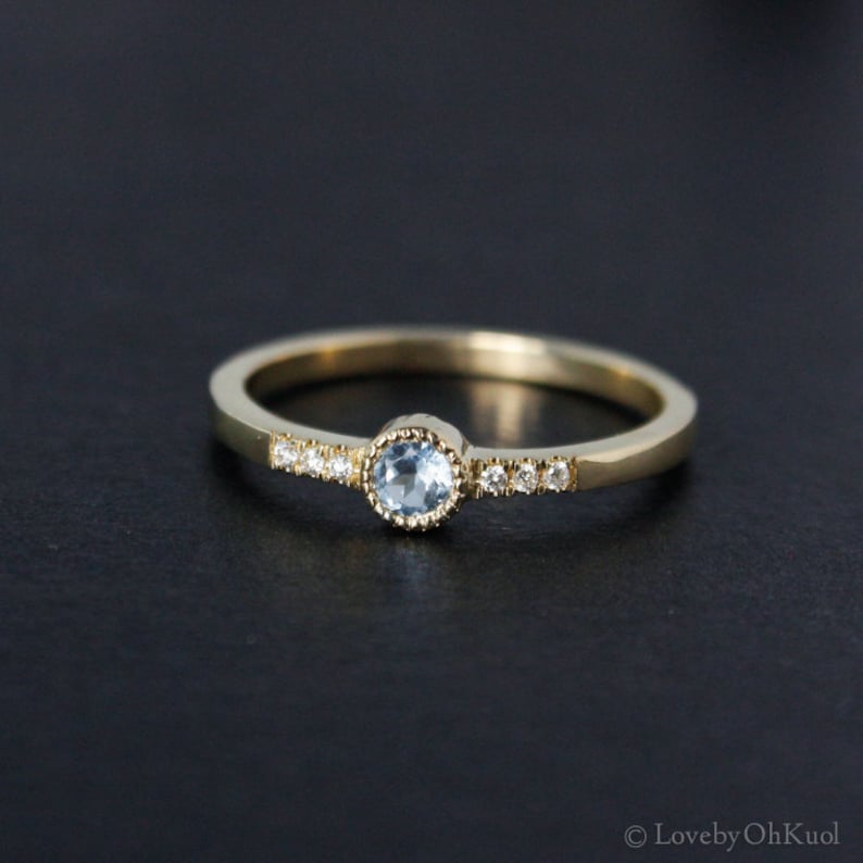 May include: A delicate gold band ring with a small, round, light blue gemstone set in a bezel setting. The band is accented with a row of tiny, clear gemstones.