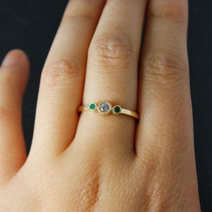 K&ouml;nnte beinhalten: Ein goldener Ring mit einem runden, klaren Stein und zwei kleineren runden gr&uuml;nen Steinen auf jeder Seite.