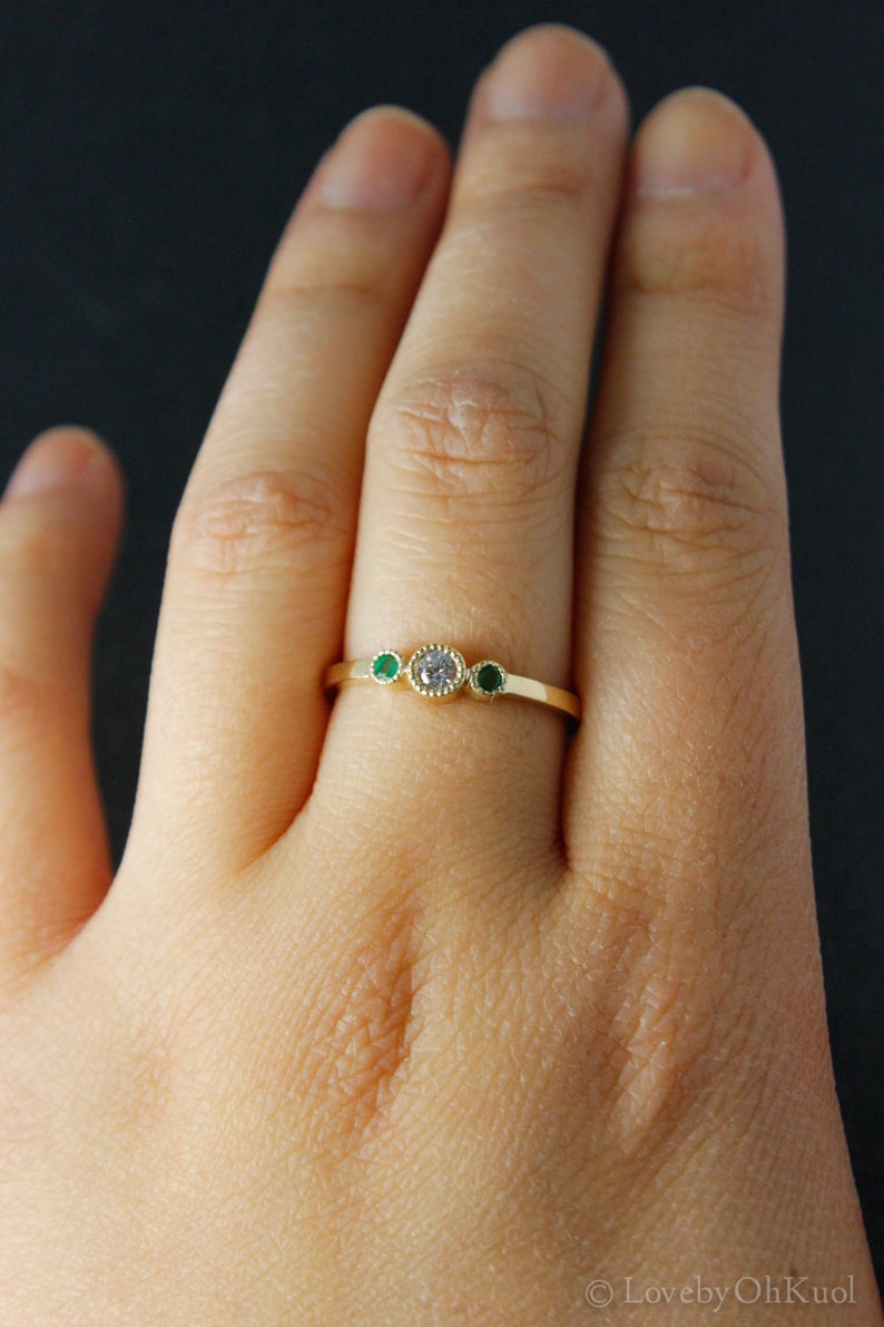 K&ouml;nnte beinhalten: Ein goldener Ring mit einem runden, klaren Stein und zwei kleineren runden gr&uuml;nen Steinen auf jeder Seite.
