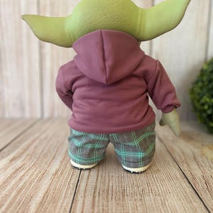 Op de afbeelding: Een groenhuidig wezenfiguur met grote puntige oren, gekleed in een mauve hoodie en een groen met zwart geruite broek. Het figuur is van achteren bekeken, met zicht op de capuchon, de lange mouwen en de zoom van de broek met patroon.
