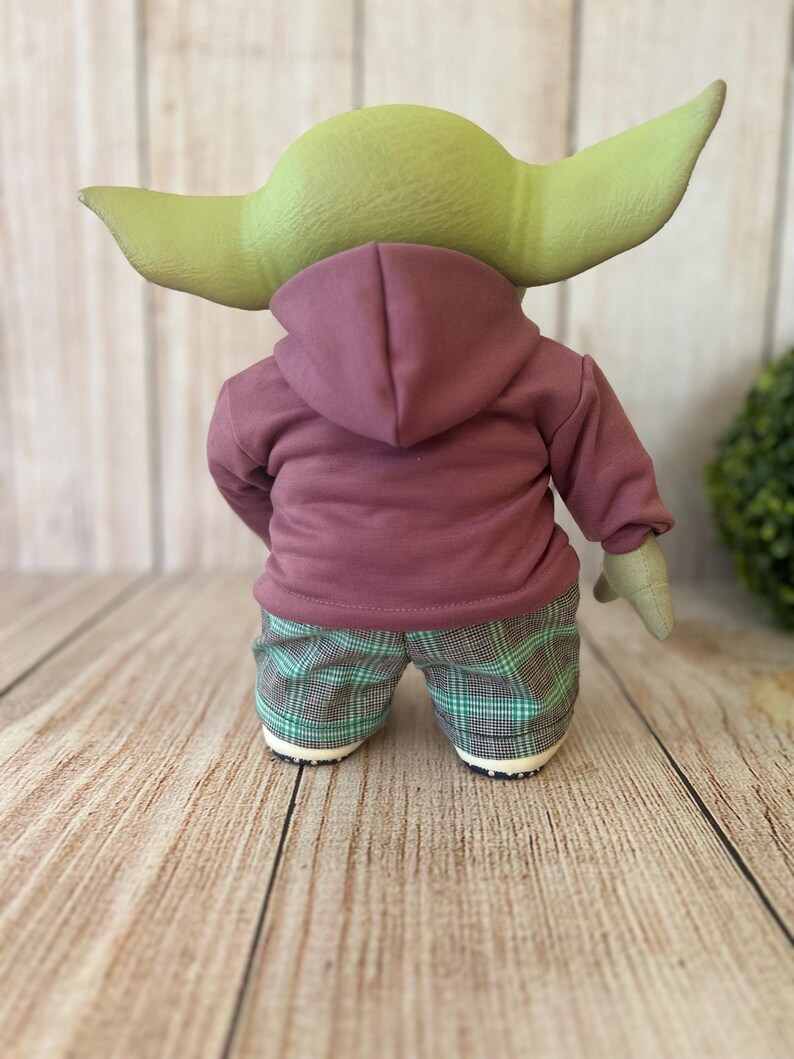 Op de afbeelding: Een groenhuidig wezenfiguur met grote puntige oren, gekleed in een mauve hoodie en een groen met zwart geruite broek. Het figuur is van achteren bekeken, met zicht op de capuchon, de lange mouwen en de zoom van de broek met patroon.