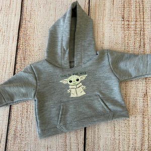 May include: A gray hooded sweatshirt featuring a graphic of a character with large ears and the text "This is the way." The sweatshirt has a kangaroo pocket and is made of a soft material.