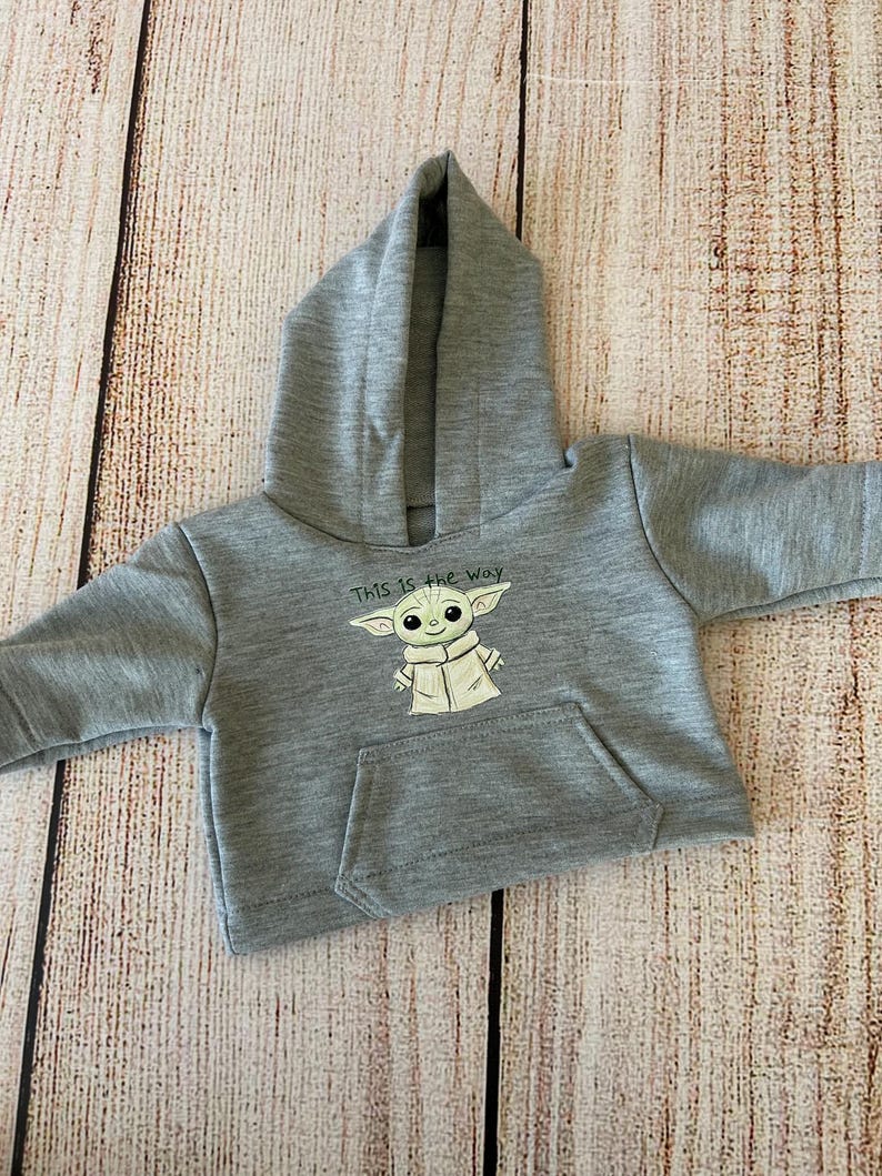 May include: A gray hooded sweatshirt featuring a graphic of a character with large ears and the text "This is the way." The sweatshirt has a kangaroo pocket and is made of a soft material.