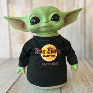 Peut inclure: Poup&eacute;e &agrave; la peau verte avec de grands yeux noirs et des oreilles pointues, portant un t-shirt noir &agrave; manches longues. Le t-shirt pr&eacute;sente un logo circulaire jaune et rouge avec les mots "Mos Eisley Cantina" et "Tatooine". La poup&eacute;e est pos&eacute;e sur une surface en bois clair.