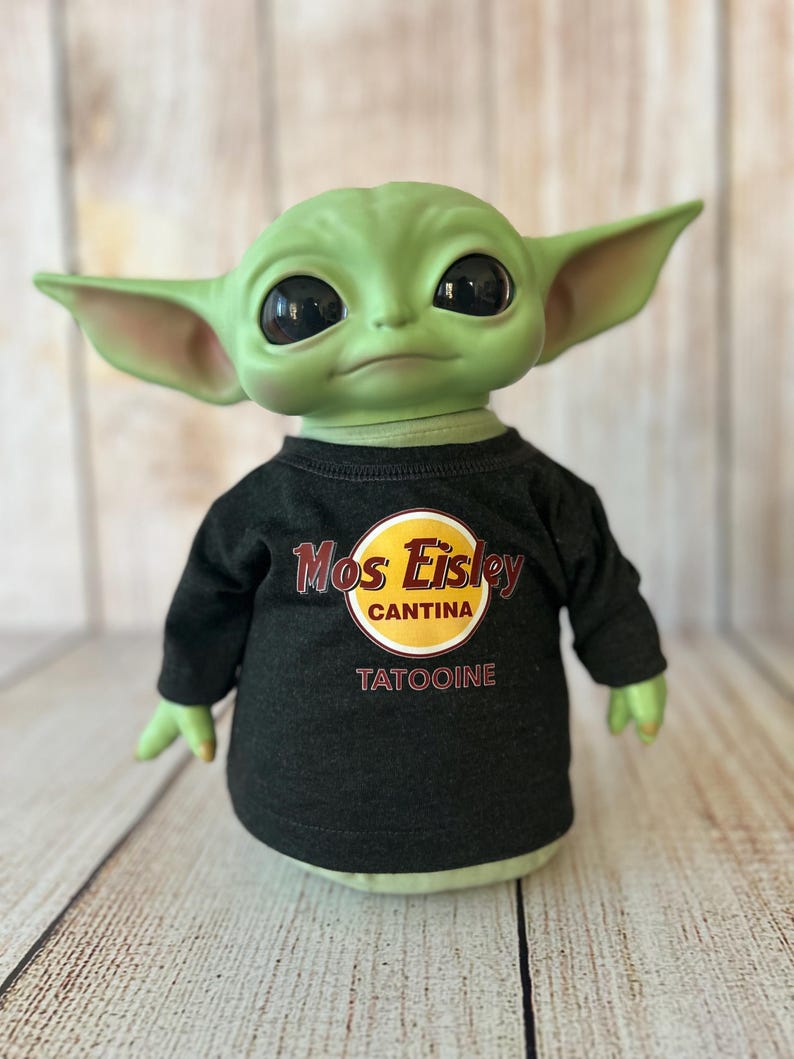 Peut inclure: Poup&eacute;e &agrave; la peau verte avec de grands yeux noirs et des oreilles pointues, portant un t-shirt noir &agrave; manches longues. Le t-shirt pr&eacute;sente un logo circulaire jaune et rouge avec les mots "Mos Eisley Cantina" et "Tatooine". La poup&eacute;e est pos&eacute;e sur une surface en bois clair.