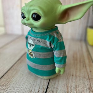 Peut inclure: Une figurine verte d'un personnage d'une s&eacute;rie de science-fiction populaire, portant un t-shirt ray&eacute; turquoise et gris. Le t-shirt porte l'inscription "This is the Way" et un dessin d'un casque.