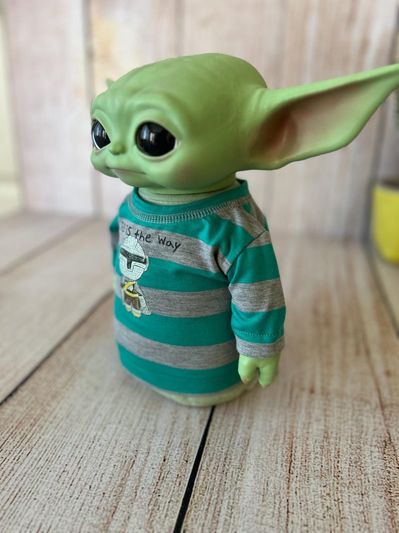 Peut inclure: Une figurine verte d'un personnage d'une s&eacute;rie de science-fiction populaire, portant un t-shirt ray&eacute; turquoise et gris. Le t-shirt porte l'inscription "This is the Way" et un dessin d'un casque.