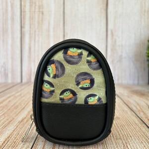 May include: A small, oval-shaped black zippered pouch featuring a light green fabric panel printed with a repeating pattern of the character Grogu from Star Wars inside a floating pod, accented by the Star Wars logo text in the background.