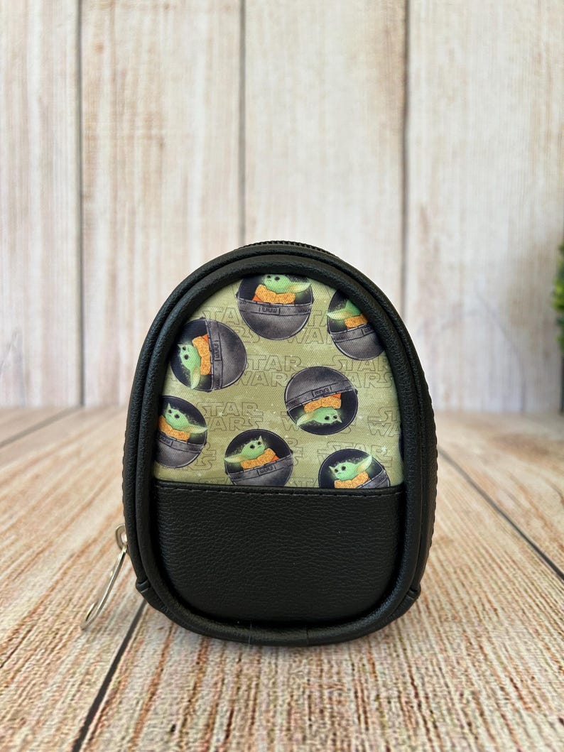 May include: A small, oval-shaped black zippered pouch featuring a light green fabric panel printed with a repeating pattern of the character Grogu from Star Wars inside a floating pod, accented by the Star Wars logo text in the background.