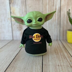 Peut inclure: Une figurine verte avec de grandes oreilles et des yeux noirs, portant un t-shirt noir &agrave; manches longues. Le t-shirt pr&eacute;sente un logo jaune et rouge qui se lit "Mos Eisley Cantina" et le mot "Tatooine" en dessous. La figurine est pos&eacute;e sur une surface en bois clair.