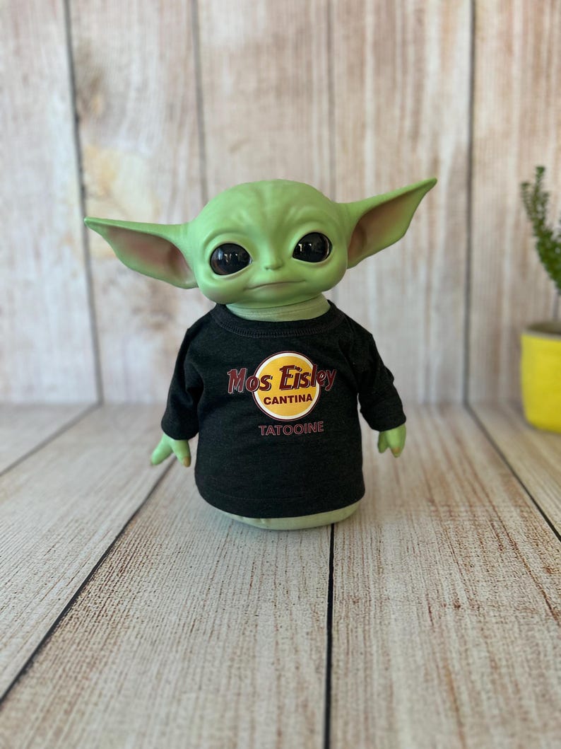 Peut inclure: Une figurine verte avec de grandes oreilles et des yeux noirs, portant un t-shirt noir &agrave; manches longues. Le t-shirt pr&eacute;sente un logo jaune et rouge qui se lit "Mos Eisley Cantina" et le mot "Tatooine" en dessous. La figurine est pos&eacute;e sur une surface en bois clair.