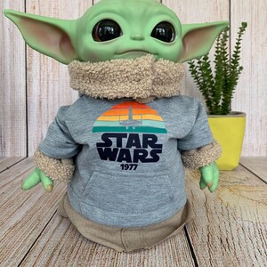 May include: A green-skinned doll with large black eyes and pointed ears, wearing a gray Star Wars hoodie with a sunset graphic and the text "STAR WARS 1977". The doll has a tan fur-lined collar and cuffs, and a tan outfit.