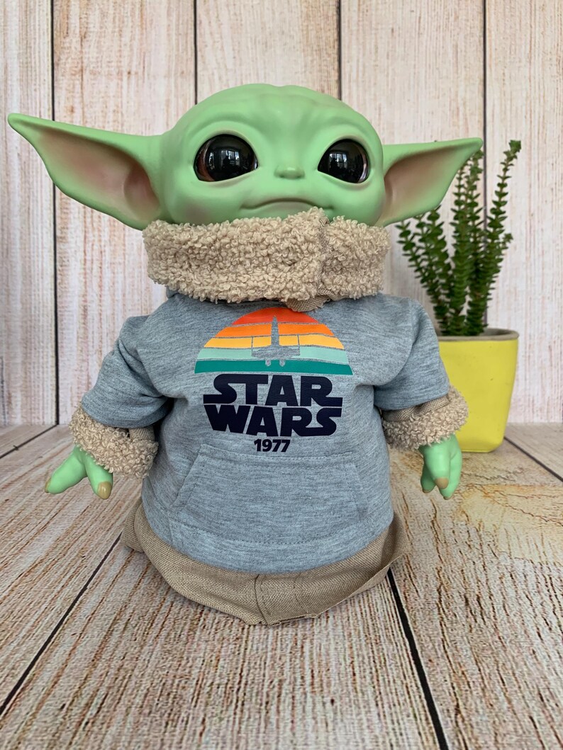 May include: A green-skinned doll with large black eyes and pointed ears, wearing a gray Star Wars hoodie with a sunset graphic and the text "STAR WARS 1977". The doll has a tan fur-lined collar and cuffs, and a tan outfit.