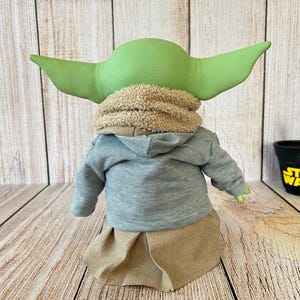 May include: A plush doll of a character with large green ears, wearing a gray hooded sweatshirt, a tan skirt, and a fuzzy scarf. The doll is posed against a wooden background.