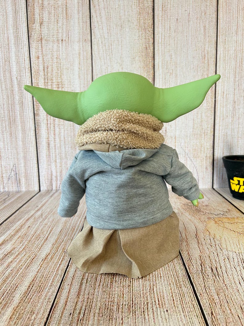May include: A plush doll of a character with large green ears, wearing a gray hooded sweatshirt, a tan skirt, and a fuzzy scarf. The doll is posed against a wooden background.