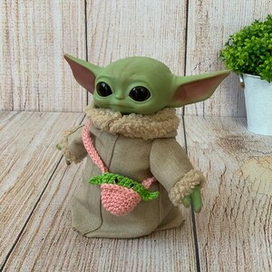 K&ouml;nnte beinhalten: Ein gr&uuml;nes Pl&uuml;schtier mit gro&szlig;en Augen und einer pinkfarbenen und gr&uuml;nen H&auml;kel-Tasche. Das Spielzeug tr&auml;gt einen braunen Mantel mit einem wei&szlig;en Pelzkragen.