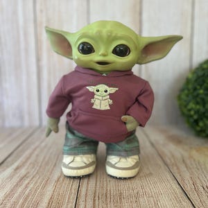 Op de afbeelding: Een groene wezenpop met grote zwarte ogen en puntige oren, gekleed in een mauve hoodie met een afbeelding van hetzelfde wezen en de tekst This is the Way, gecombineerd met een groene geruite broek en witte sneakers met grijze accenten.