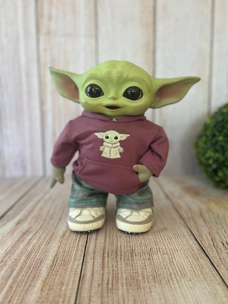 Op de afbeelding: Een groene wezenpop met grote zwarte ogen en puntige oren, gekleed in een mauve hoodie met een afbeelding van hetzelfde wezen en de tekst This is the Way, gecombineerd met een groene geruite broek en witte sneakers met grijze accenten.